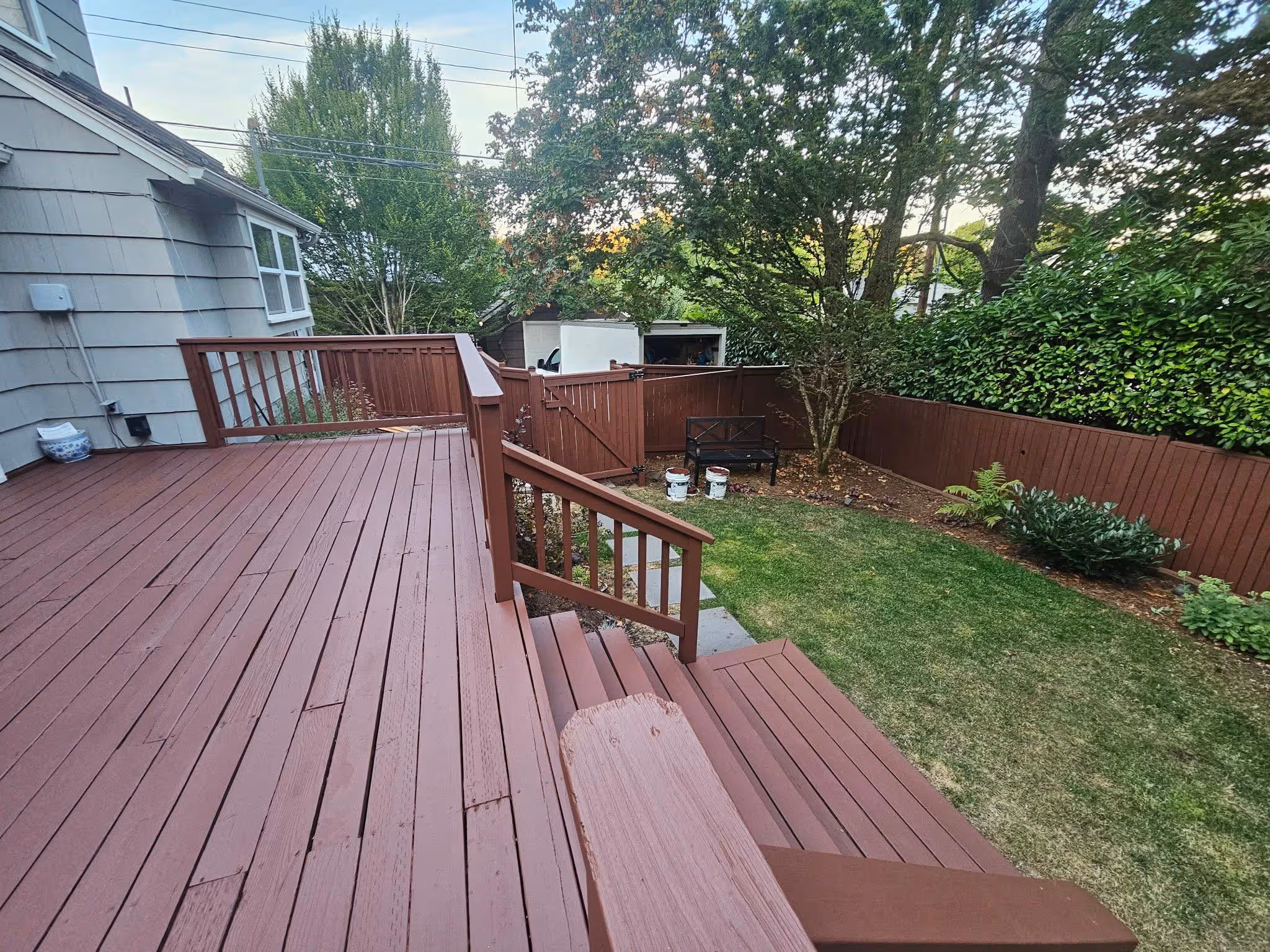 Backyard with brown wooden deck, stairs, lawn, trees, plants, and a small black bench by a fence.