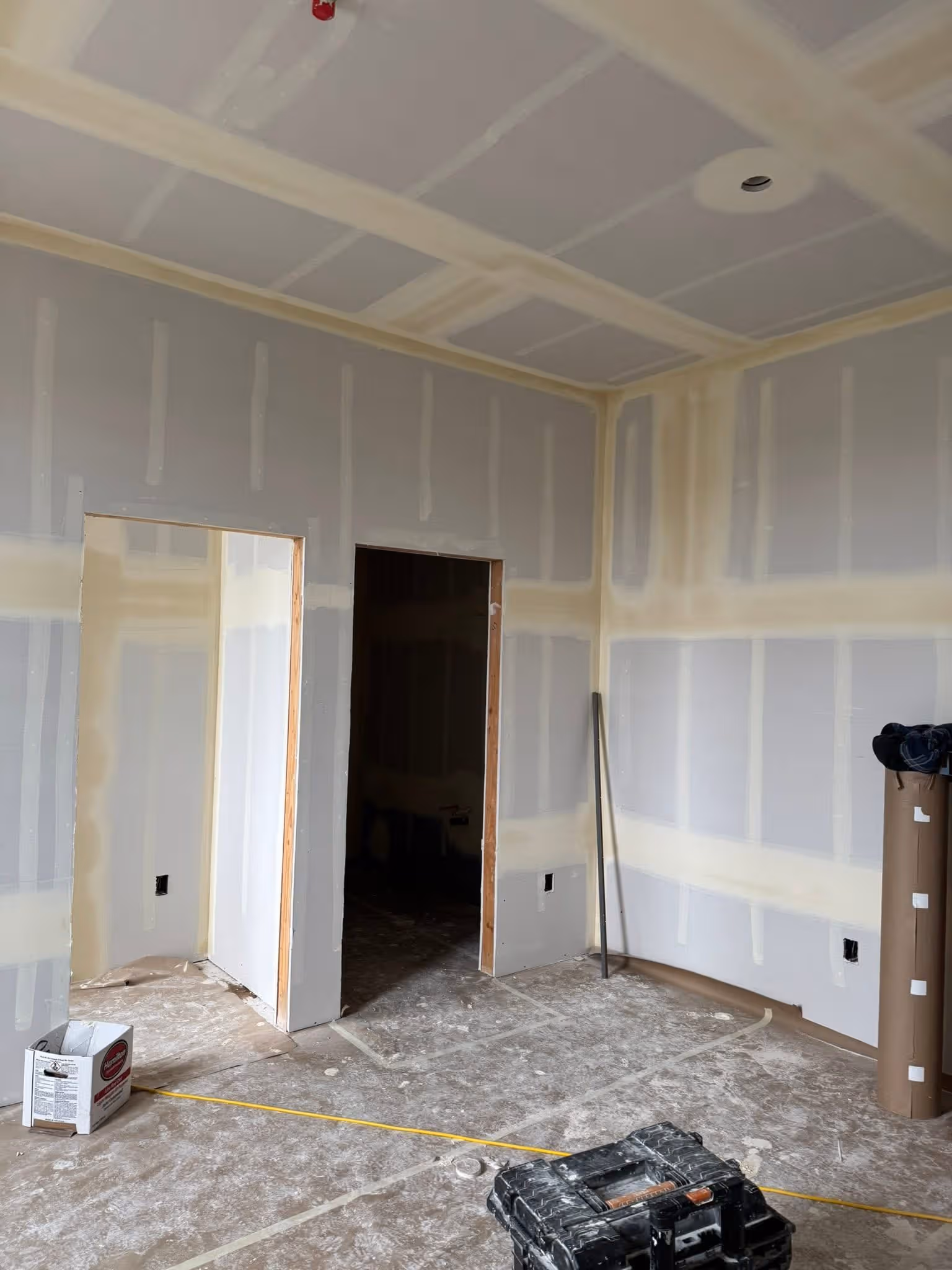 Room under construction with drywall installed, taped seams, two doorways, and construction tools on the floor.