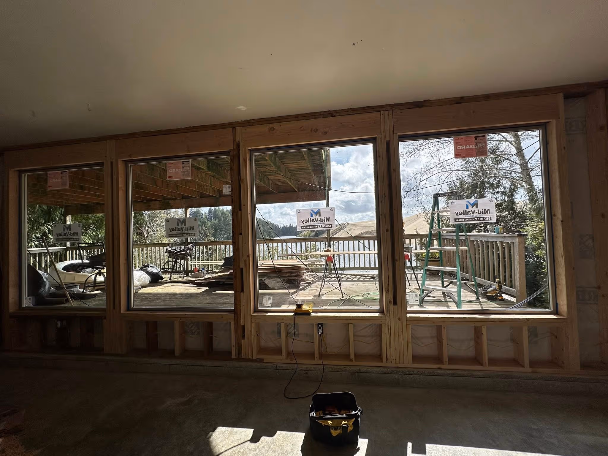 Four large framed windows under construction showing a deck with construction tools and a lake view outside.