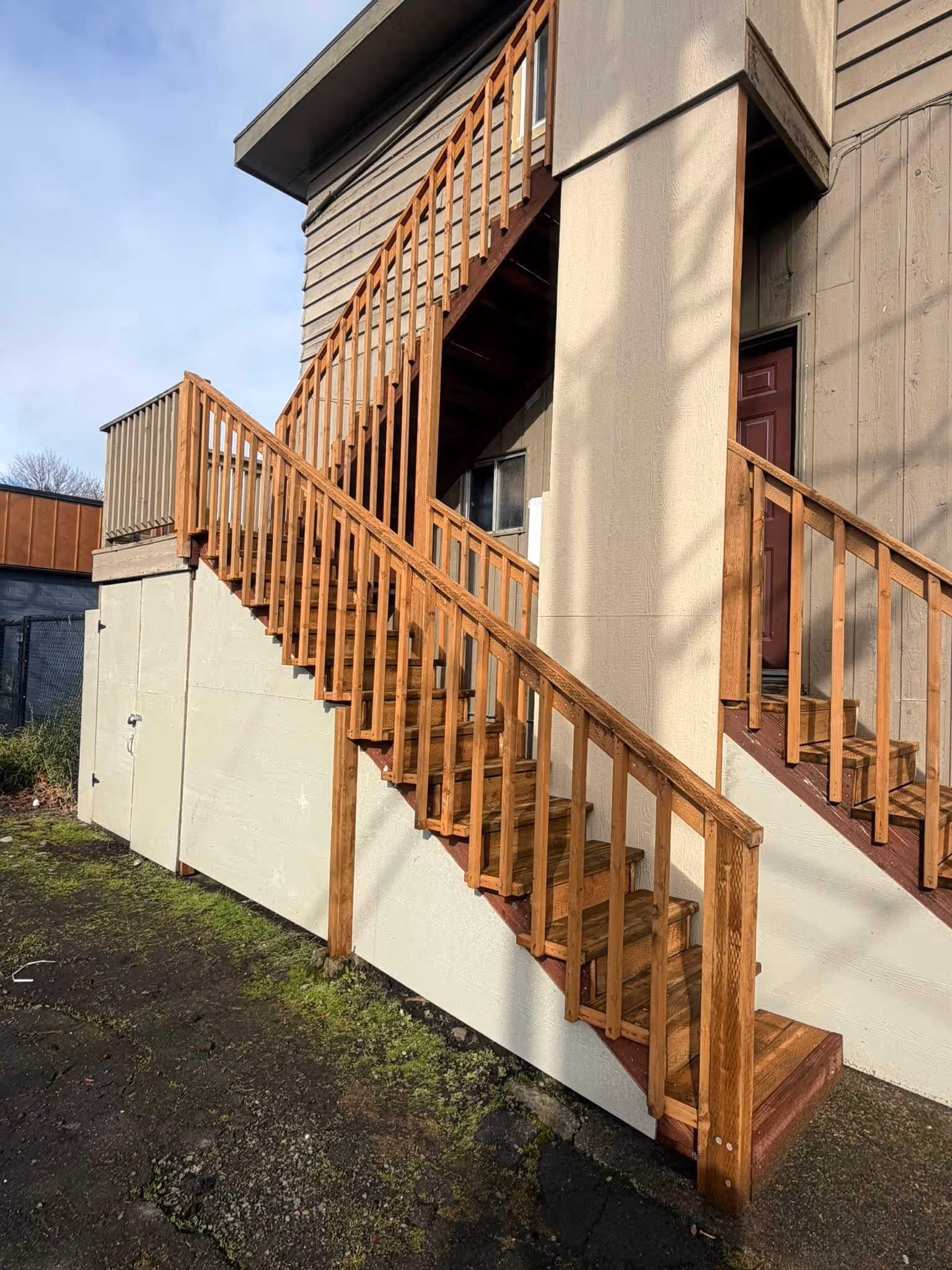 Outdoor wooden staircase with railings leading to two levels of a beige building with a red door.