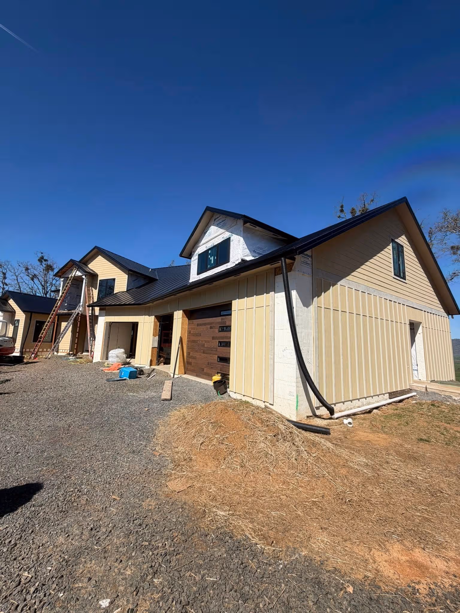 A large house under construction with yellow siding, two dormer windows, and a gravel driveway.