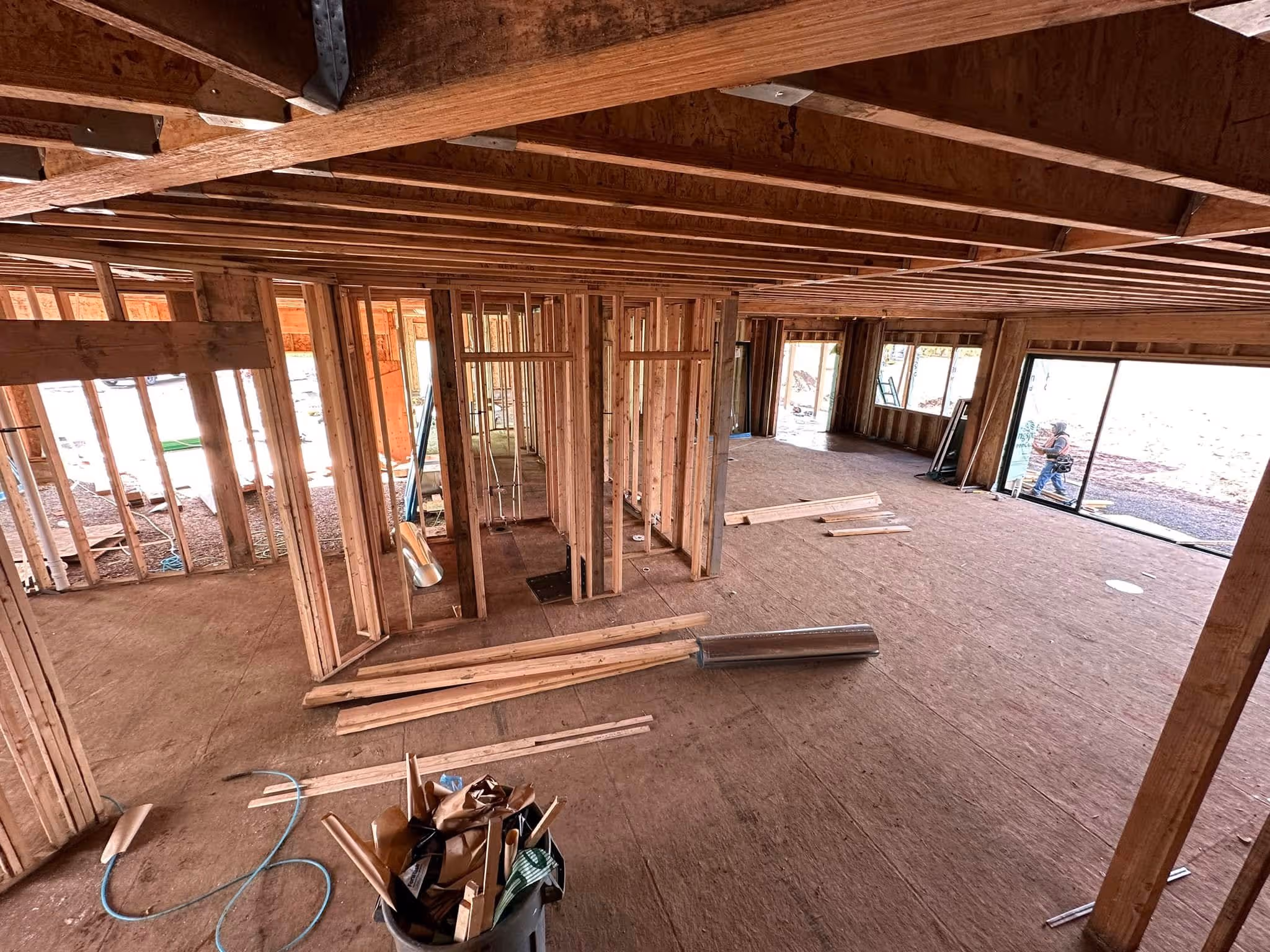 Interior of a house under construction with exposed wooden framing and a worker outside sliding glass doors.