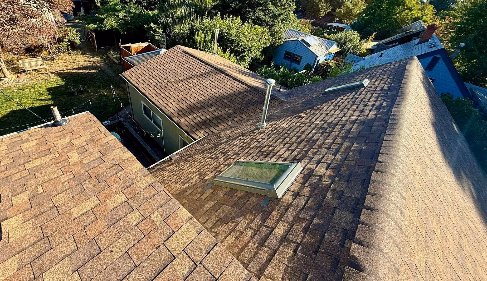 Brown shingled rooftops with skylights and chimneys overlooking a backyard with trees and houses.